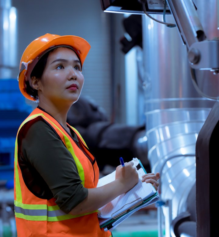 Woman in high visibility vest and hard hat with clipboard inspecting indoor factory equipment
