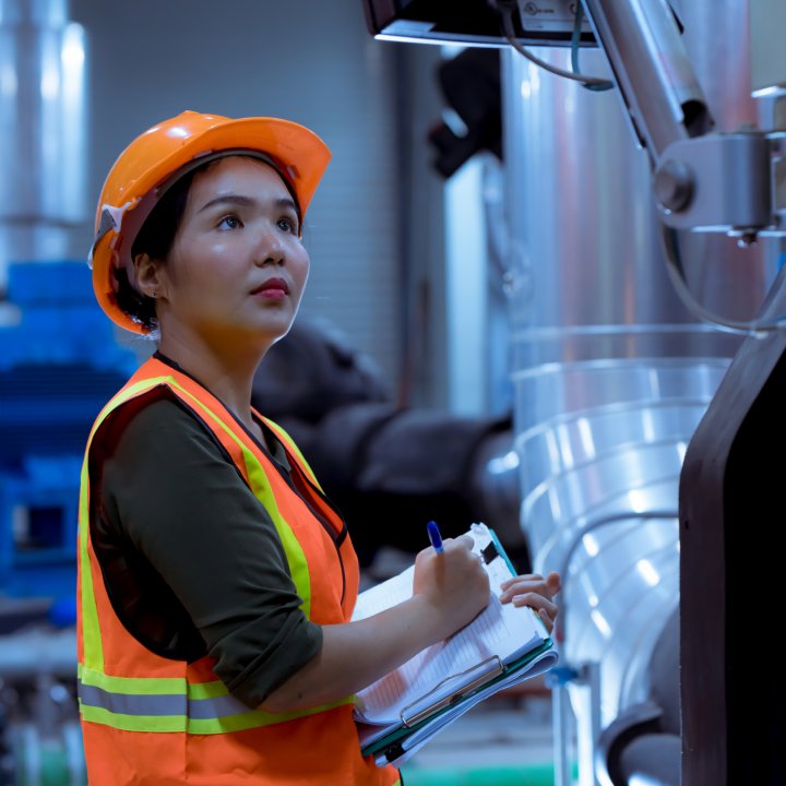 Woman in high visibility vest and hard hat with clipboard inspecting indoor factory equipment