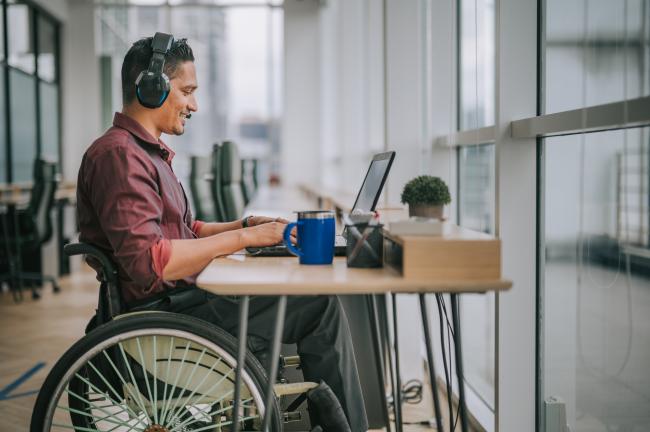 Small business customer sitting at desk in front of window, using laptop and headphones